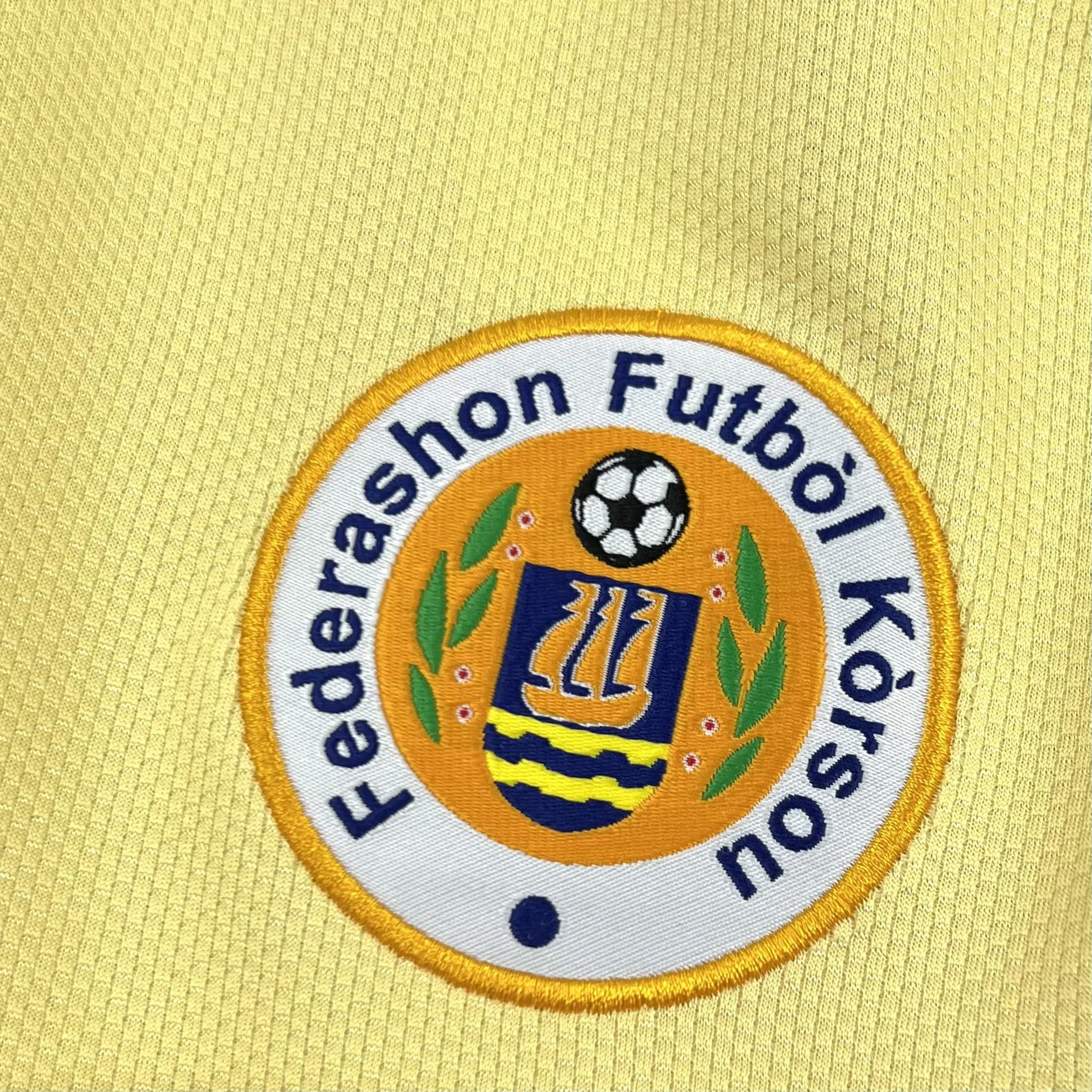 Primer plano del escudo de la Federación de Fútbol de Curazao bordado en azul sobre el tejido técnico blanco.