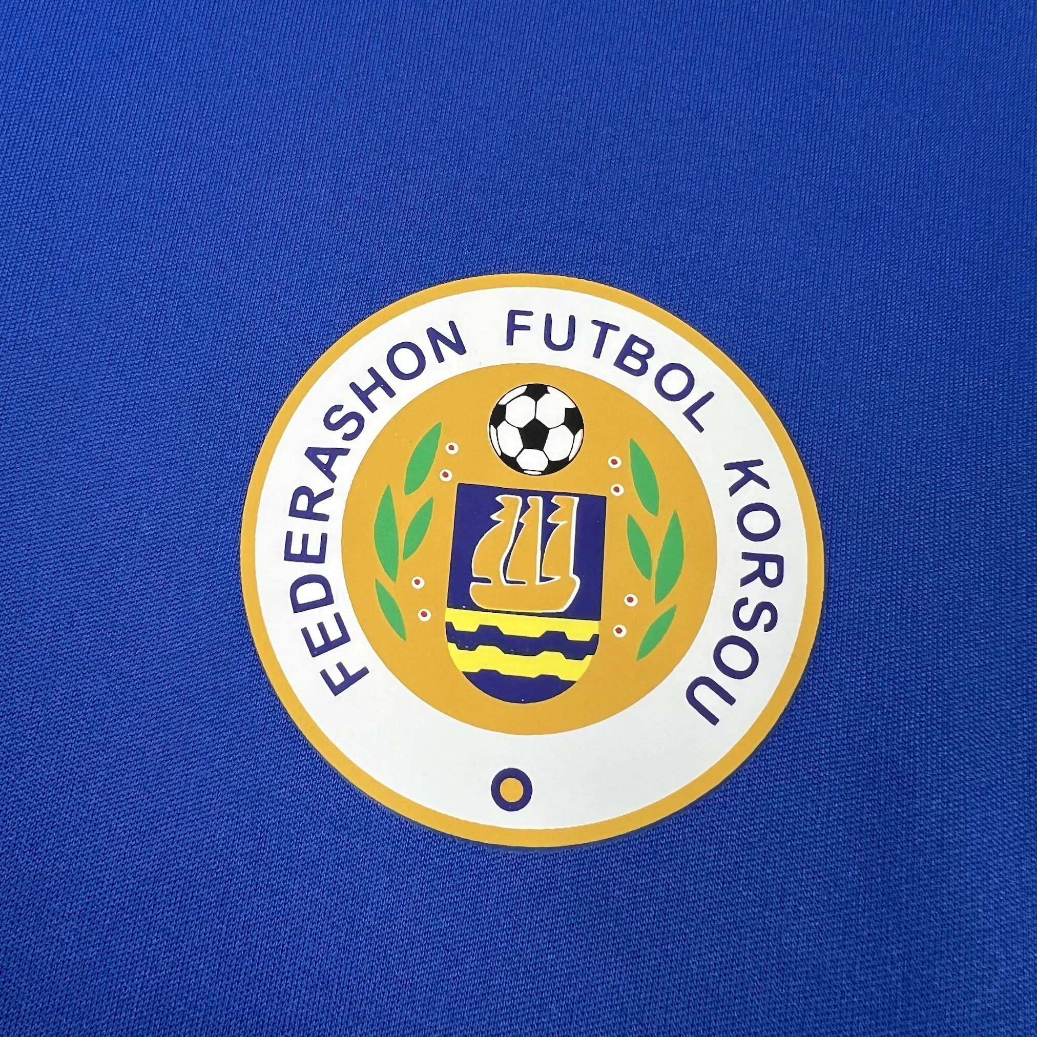 Primer plano del escudo de la Federación de Fútbol de Curazao bordado sobre el tejido azul texturizado.
