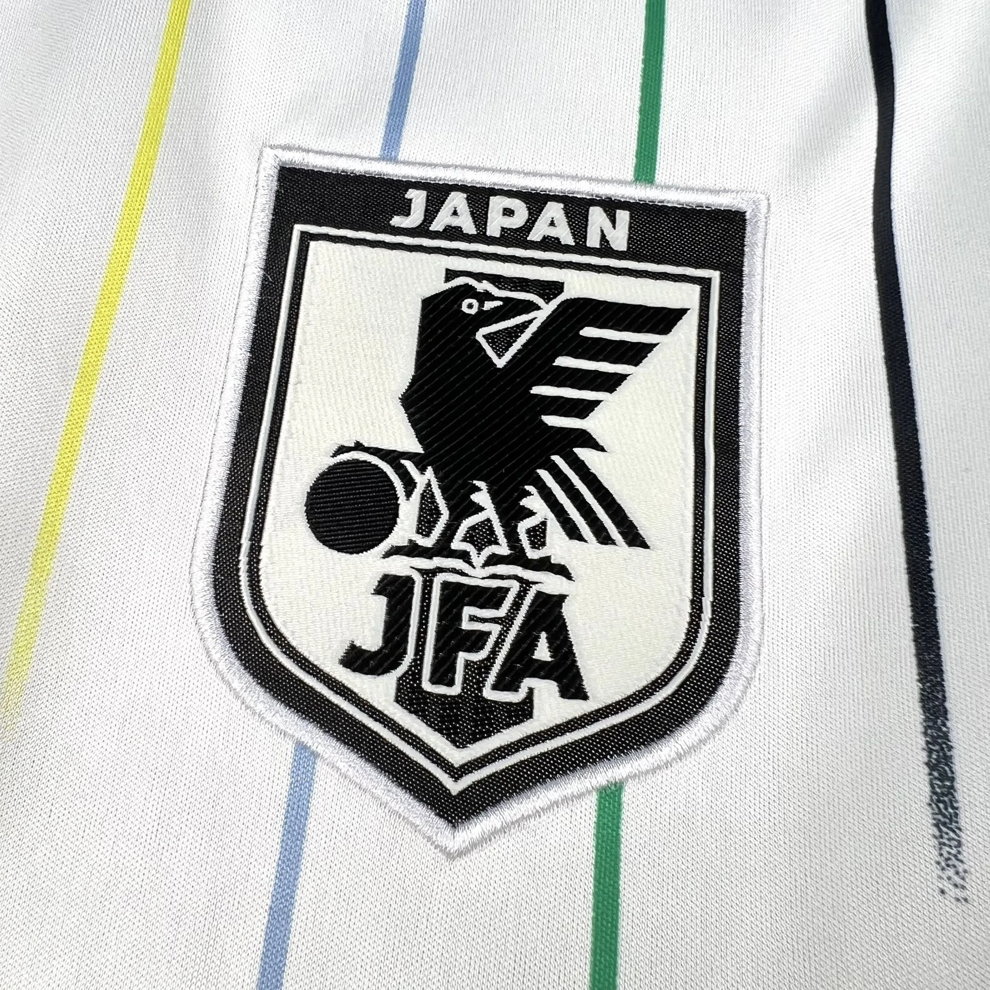 Primer plano del escudo de la selección japonesa de fútbol (JFA) bordado sobre el tejido blanco texturizado.
