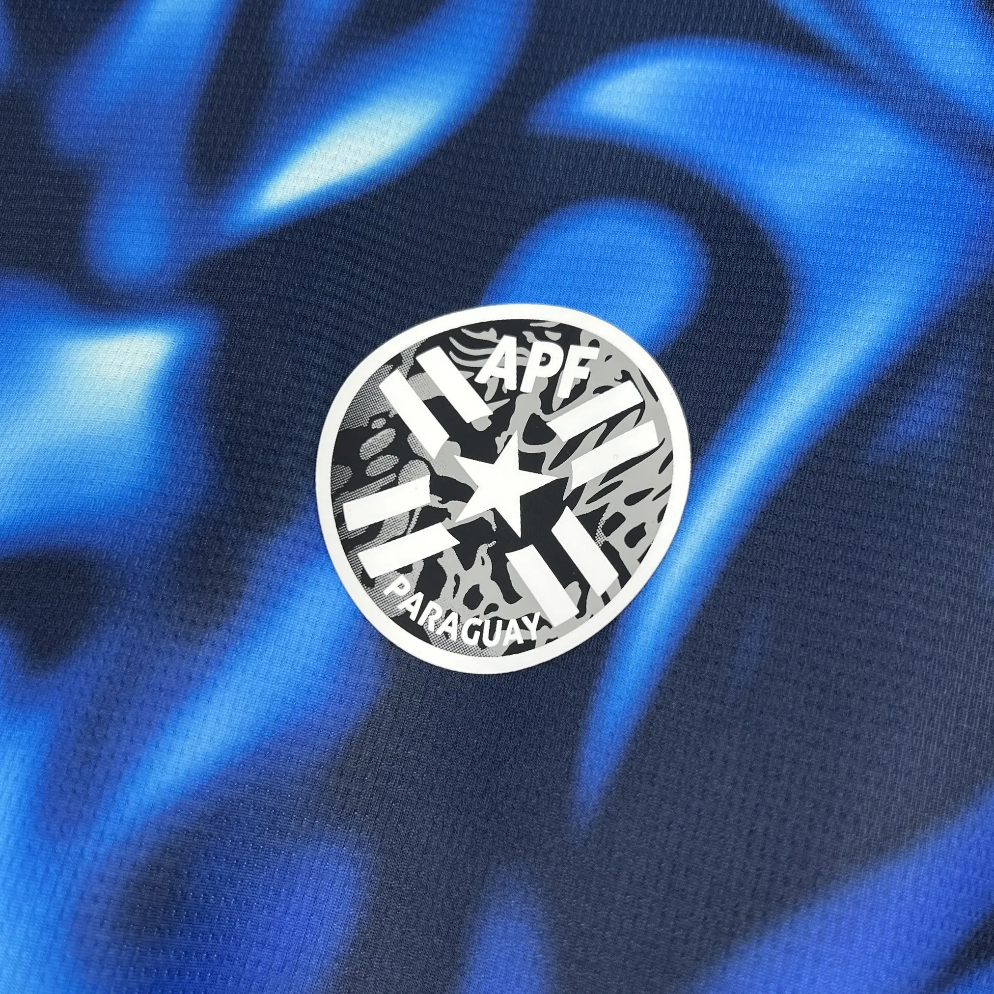 Primer plano del escudo de la selección de Paraguay bordado sobre el fondo azul con detalles de ondas gráficas.