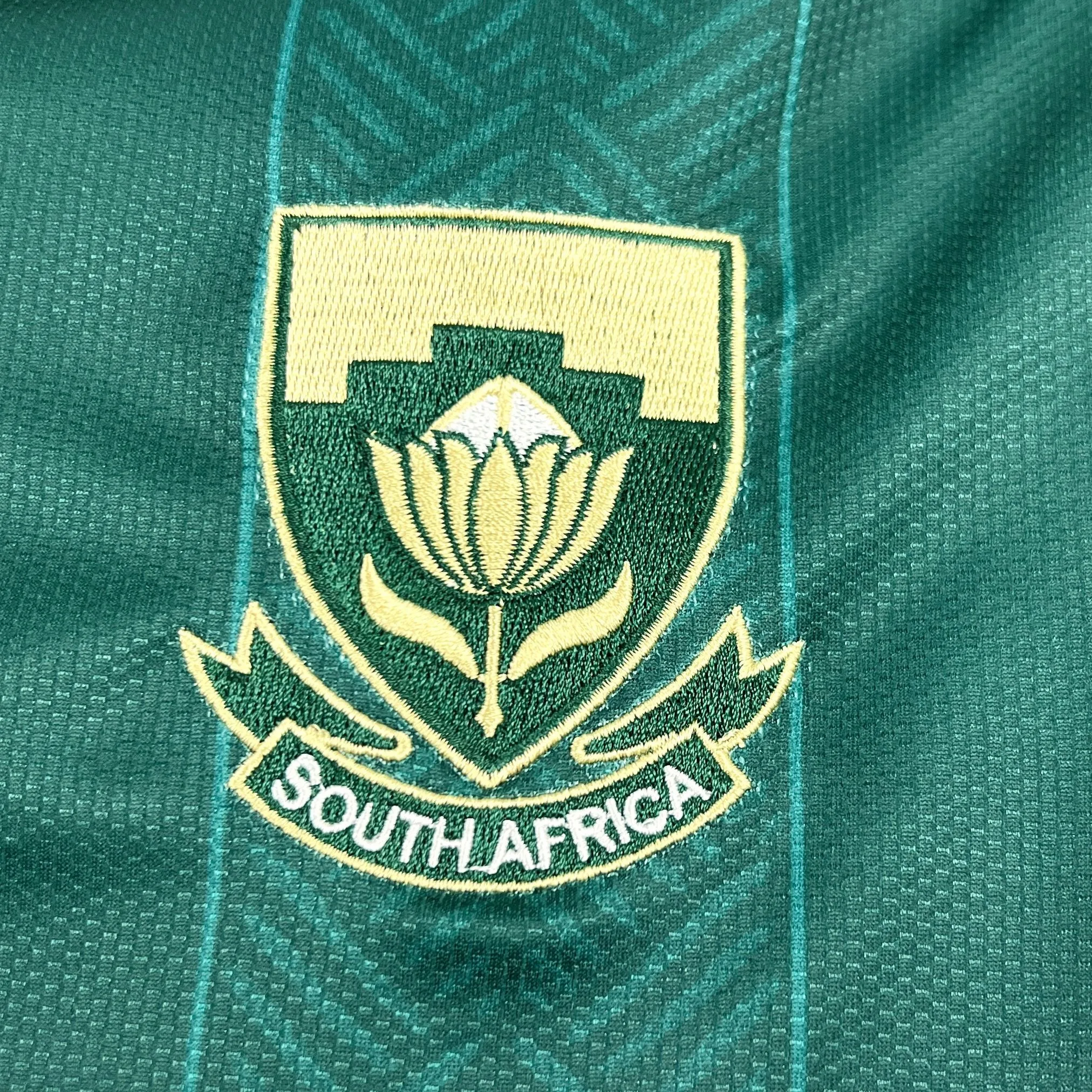 Detalle del parche de la flor nacional Protea bordado en color amarillo sobre el fondo verde de la prenda.