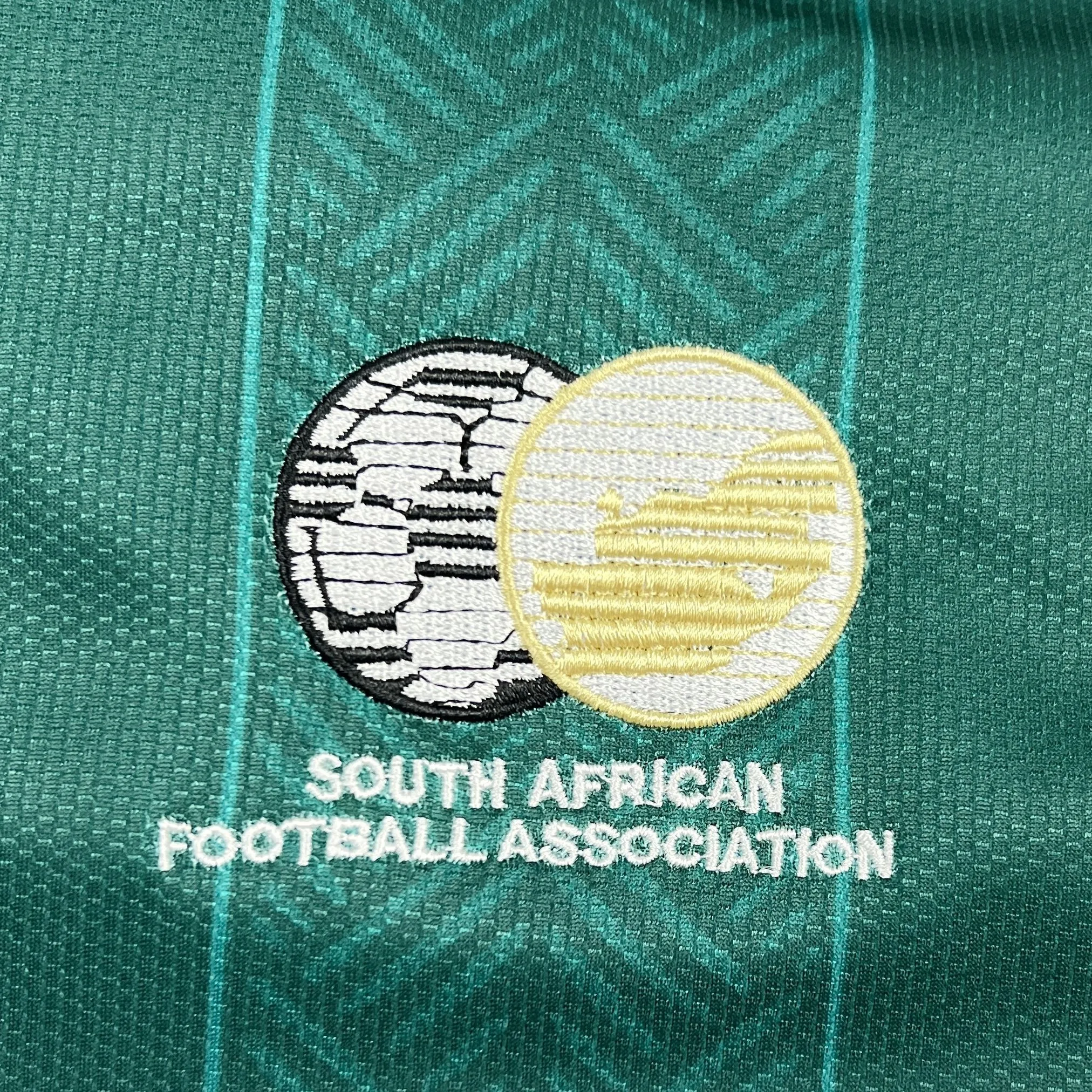 Primer plano del escudo de la Asociación de Fútbol de Sudáfrica (SAFA) bordado en la parte frontal de la camiseta.