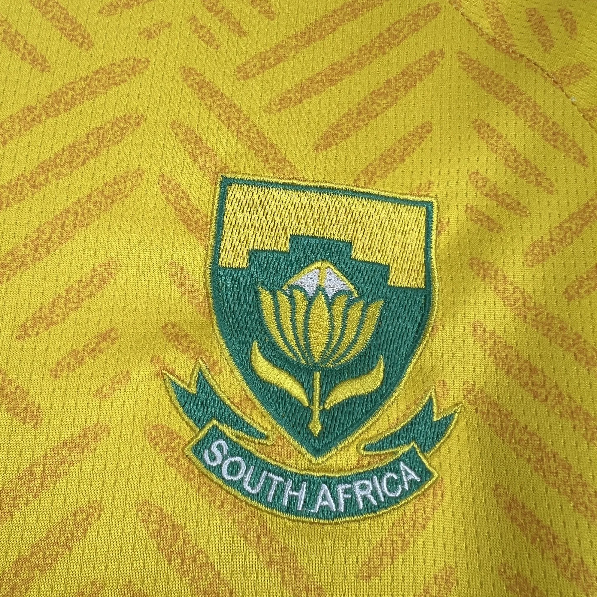 Detalle del parche de la flor nacional de Sudáfrica, la Protea, bordado en color verde sobre el pecho de la camiseta.