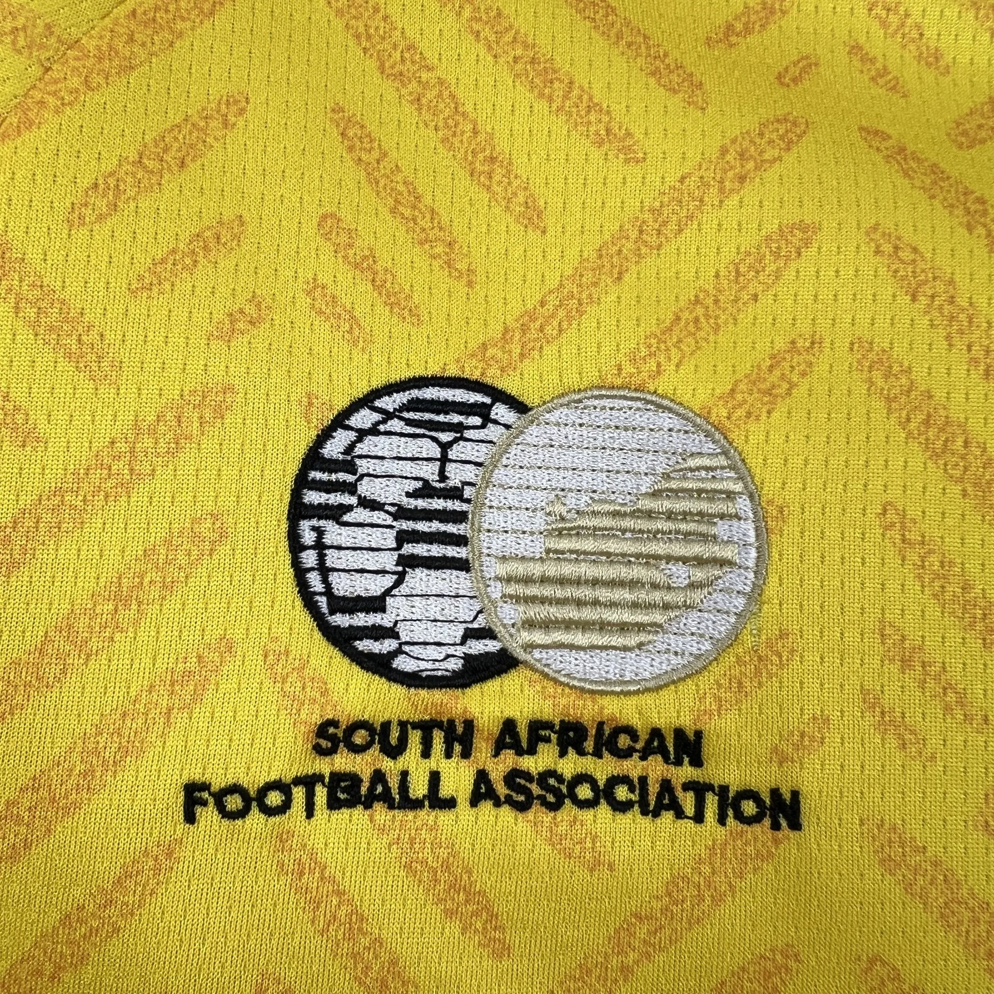 Primer plano del escudo de la Asociación de Fútbol de Sudáfrica (SAFA) bordado sobre el fondo amarillo de la prenda.
