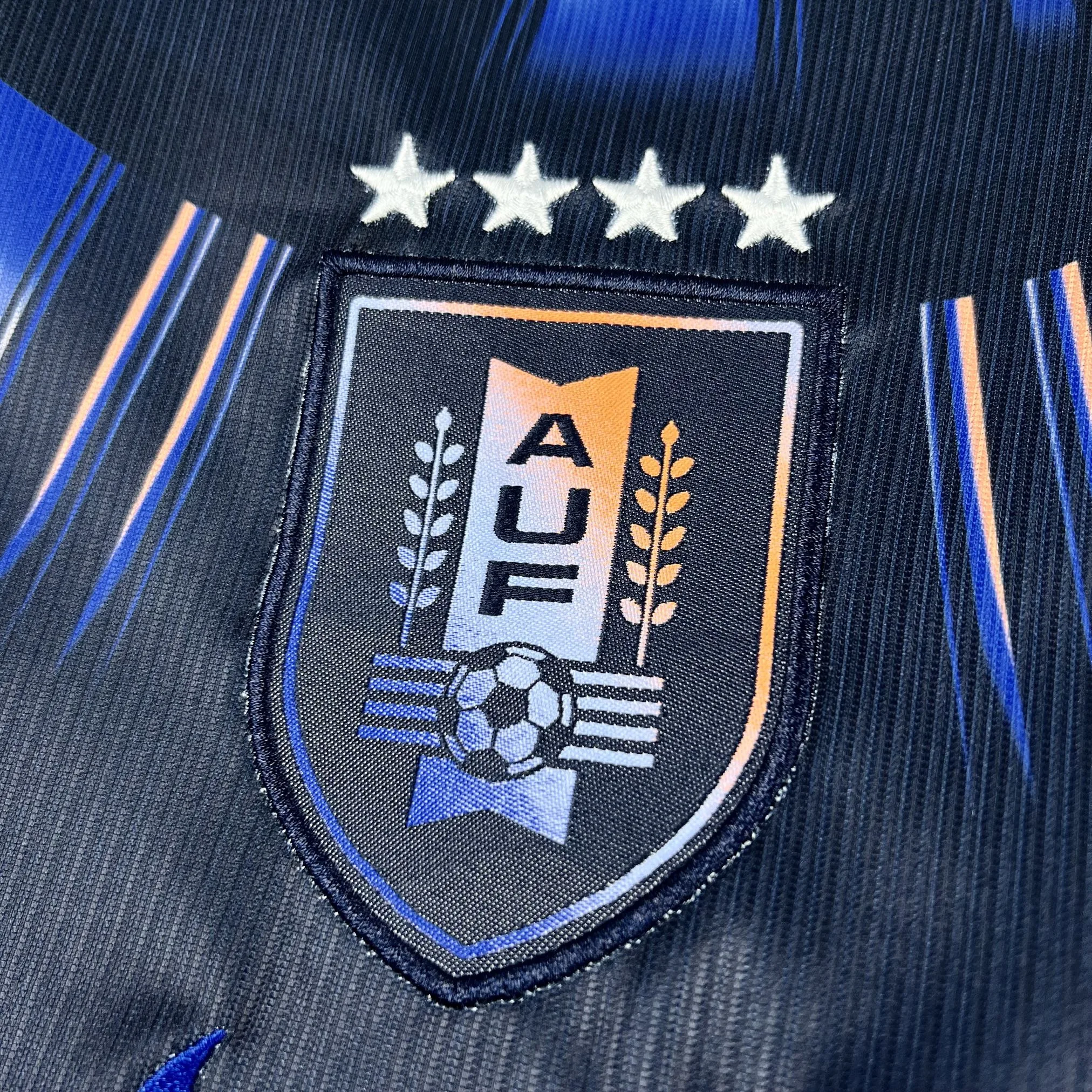 Primer plano del escudo de la Asociación Uruguaya de Fútbol con sus cuatro estrellas doradas, bordado sobre el tejido blanco.
