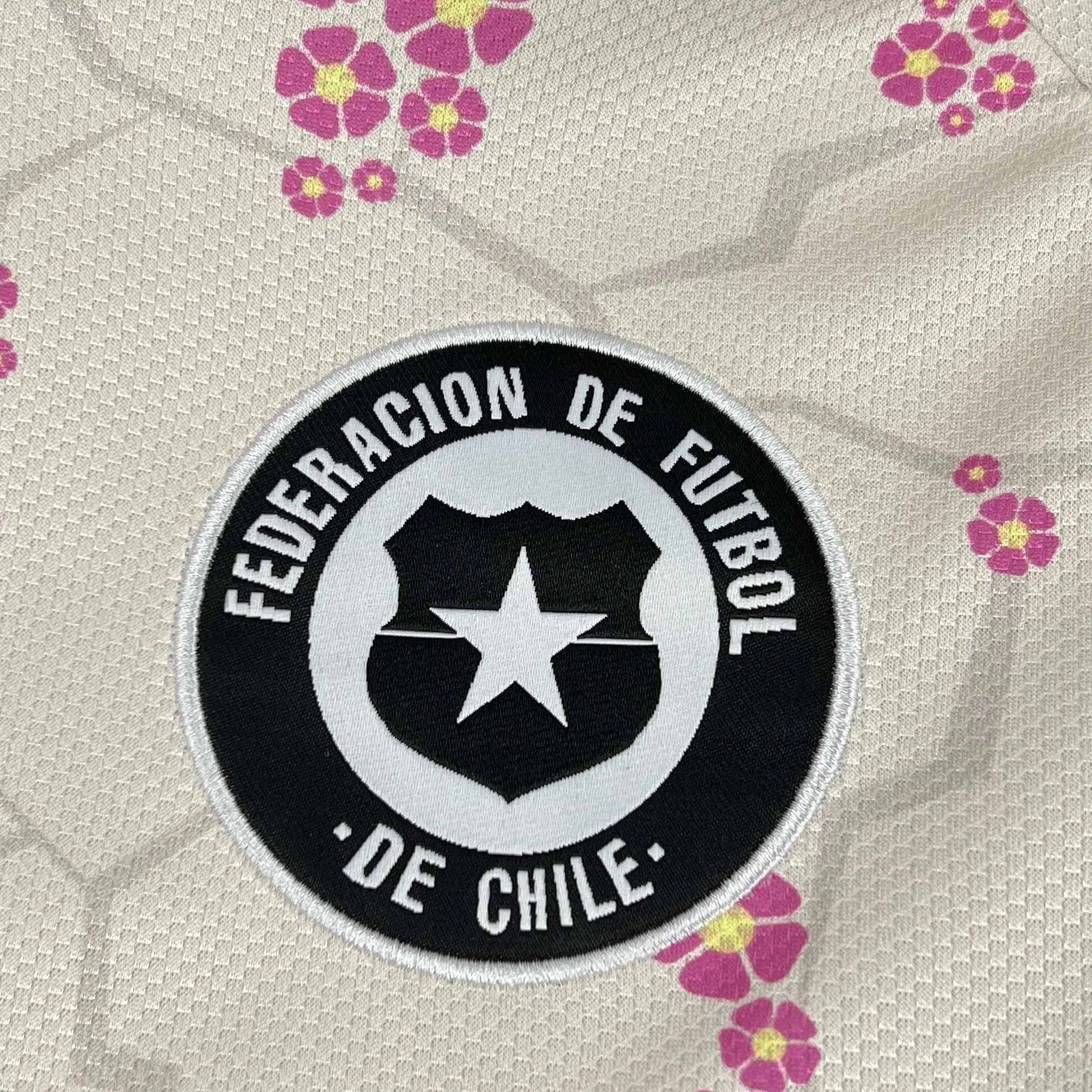 Primer plano del escudo de la Federación de Fútbol de Chile bordado en sus colores originales sobre el fondo blanco.