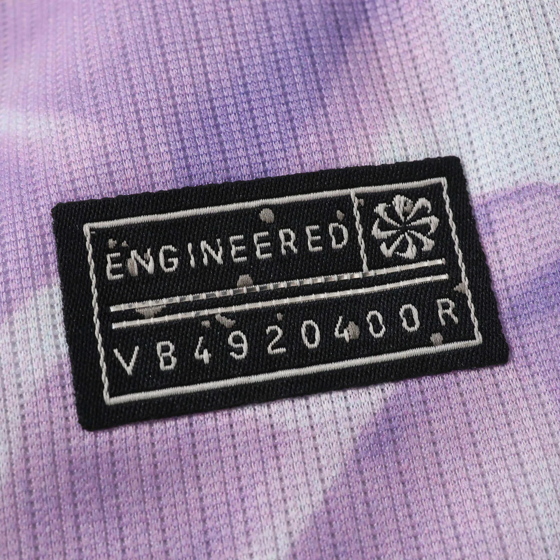 Detalle del sello rectangular de autenticidad "Engineered" sobre el tejido texturizado morado.