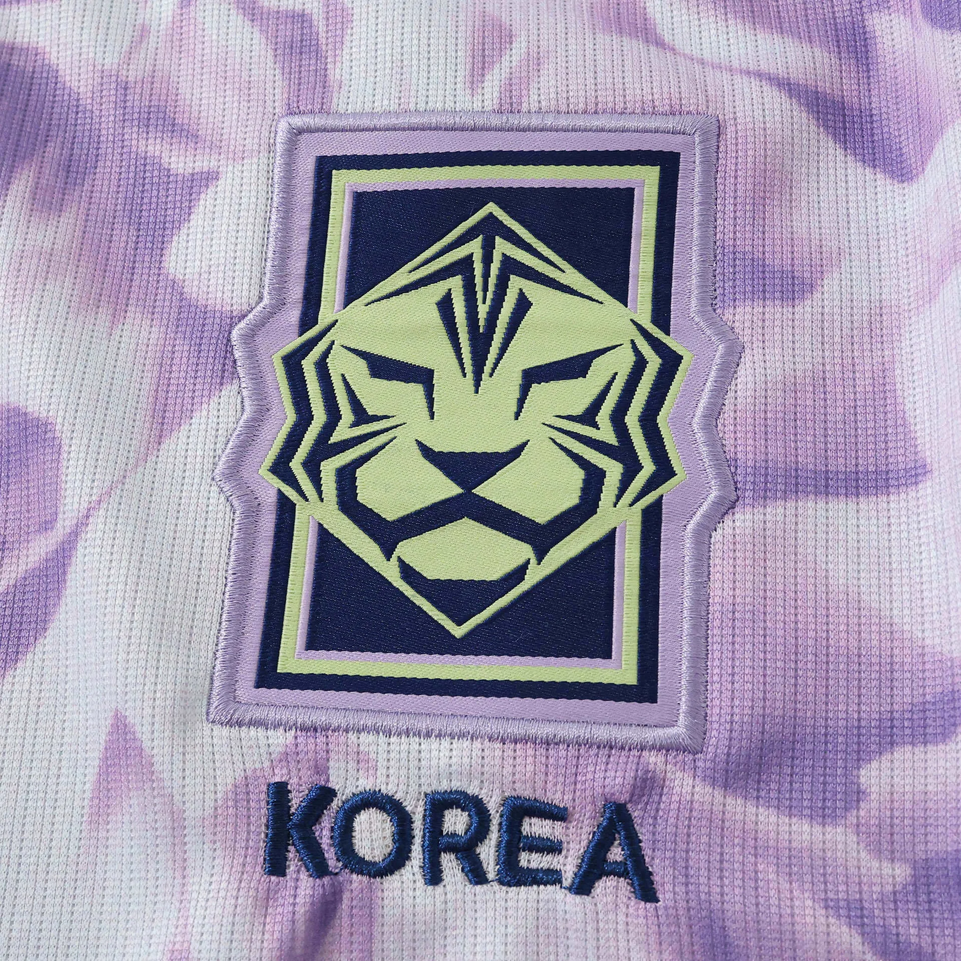 Primer plano del escudo de la selección de Corea del Sur bordado en color azul marino y crema sobre el patrón de la camiseta.