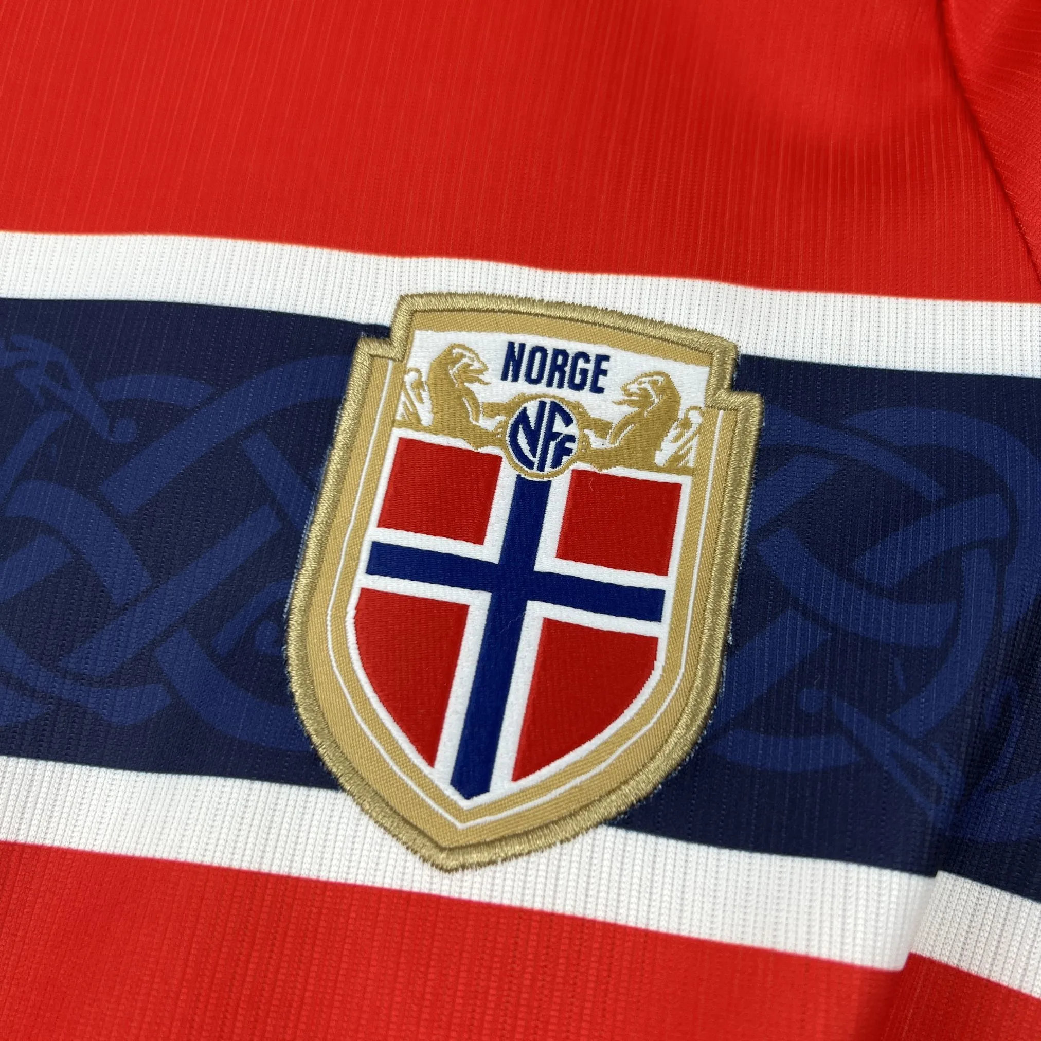 Primer plano del escudo de la selección de Noruega con la bandera nacional, bordado sobre el tejido técnico rojo.