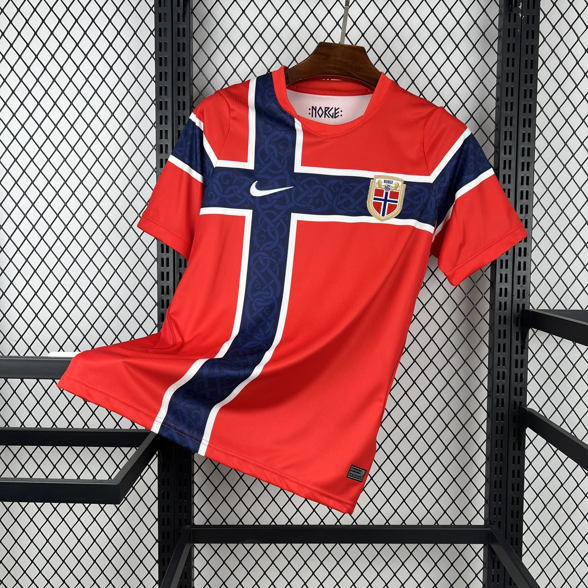 Imagen frontal completa de la camiseta local de Noruega, color rojo con una llamativa franja central azul y blanca.