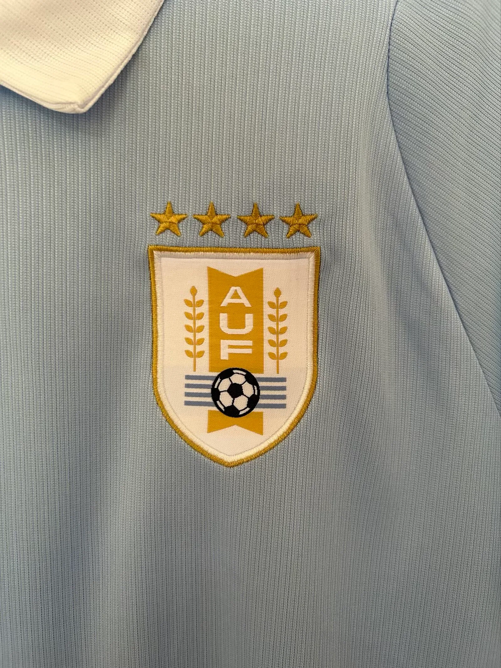 Primer plano del escudo de la Asociación Uruguaya de Fútbol con sus cuatro estrellas doradas, bordado sobre el tejido celeste.